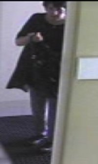 Kolejne ujęcie. Kobieta wchodząca do placówki wymiany walut, którą zarejestrowały kamery monitoringu. Ubrana w jasne spodnie, ciemną długą koszulkę. Kobieta ma ciemne, krótkie włosy. Na zdjęciu widać jak zakłada maseczkę.