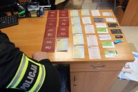 Policjant spisuje karty i dowody rejestracyjne od pojazdów i kart bankomatowe leżące na biurku.