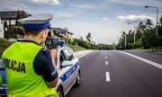 Policjant ruchu drogowego stoi na drodze i mierzy prędkość przejeżdżających pojazdów miernikiem prędkości, który trzyma w ręce