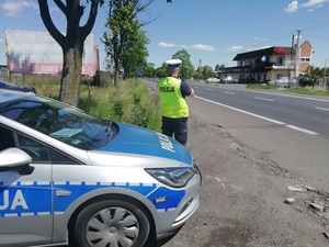 Za radiowozem stoi policjant, który mierzy prędkośćc jadących drogą samochodów