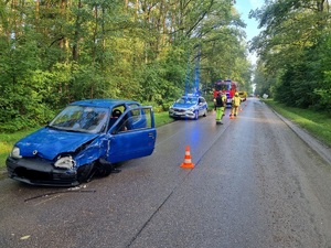 Droga asfaltowa wzdłuż lasu. Po lewej stronie drogi stoi niebieski samochód osobowy marki Fiat, ma uszkodzony przód oraz stronę kierowcy, dalej stoi radiowóz, wóz strażacki, stoją mundurowi.