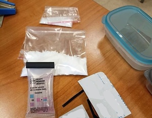 Na stole leżą dwie foliowe torebki z zawartością białego proszku, opakowania po witaminach, plastikowe przezroczyste pudełko z niebieską pokrywką oraz tester narkotykowy.