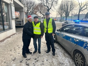 Mężczyzna ubrany w kamizelkę odblaskową, stoi obok niego policjantka w czapce mikołajowej, a z jego drugiej strony policjant.