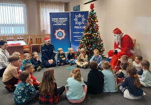 Święty Mikołaj siedzi na krzesełku po prawej stronie, dzieci siedzą na podłodze wokół niego