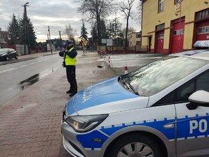 Na chodniku przy drodze stoi policjant, patrzy przez urządzenie do pomiaru prędkości, obok stoi radiowóz.