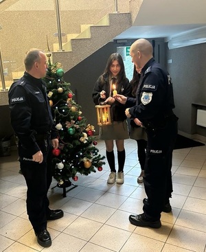 Przy choince stoją harcerki oraz policjanci. Policjant zapala od harcerek świeczkę z Betlejemskiego Światła Pokoju.