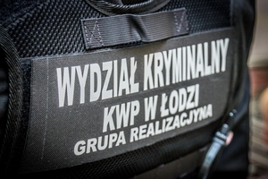 Policjant stoi tyłem, ma założona kamizelkę taktyczną, na plecach napis: Wydział Kryminalny KWP w Łodzi Grupa Realizacyjna
