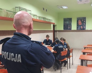 Uczniowie siedzą przy stole, rozwiązują wspólnie test, policjant stoi w pobliżu, przygląda się im.