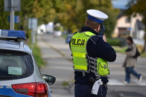 Tyłem stoi umundurowany policjant ruchu drogowego, który urządzeniem do kontroli prędkości sprawdza, jak szybko kierowca, który zbliża się do przejścia dla pieszych. Na przejściu idzie pieszy.