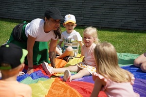Na kolorowym materiale, na trawniku siedzą dzieci, jest z nimi kobieta, rozmawia z dziećmi.