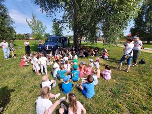 Dzieci siedzą w cieniu pod drzewem, przed nimi stoi policjantka, mówi do nich.