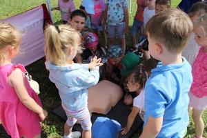 Dzieci są zebrane wokół ratowniczki, ćwiczą resuscytację krążeniowo-oddechową na fantomie.