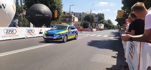 Trasa przejazdu, wzdłuż niej banery reklamowe, balony, publiczność, środkiem ulicy jedzie radiowóz policyjny.