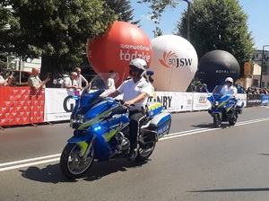 Trasa przejazdu, wzdłuż niej banery reklamowe, balony, publiczność, środkiem ulicy jedzie dwóch policjantów na służbowych motocyklach.