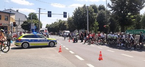 Skrzyżowanie w mieście, stoi na nim radiowóz policyjny. Wokół chodzą kolarze z rowerami, zgromadzona jest publiczność