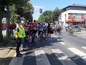 Skrzyżowanie miasta, stoi radiowóz, po lewej stronie stoi umundurowana policjantka, wokół chodzi dużo osób, uczestników wyścigu, są też kolarze z rowerami.