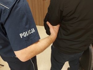 Umundurowany policjant po lewej stronie zdjęcia, po prawej zatrzymany, policjant trzyma go za przedramię.