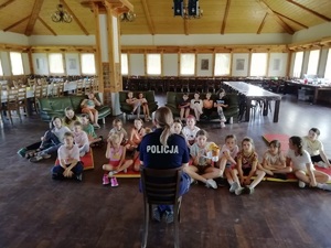 Policjantka siedzi na krzesełku tyłem do zdjęcia, ale przodem do dzieci, które siedzą na podłodze przed nią.