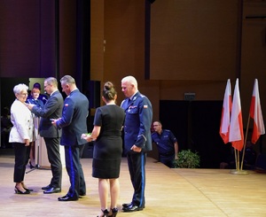 Na scenie, po lewej stronie stoi kobieta, stojący przed nią mężczyzna wpina jej medal, obok stoją umundurowani policjanci.