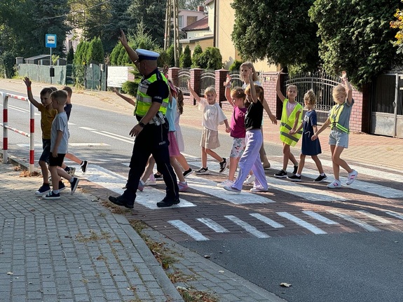 Umundurowany policjant w odblaskowej kamizelce przeprowadzający dzieci przez przejście dla pieszych.