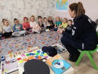 na pierwszym planie widoczna st.sierż. Kamila Sowińska , która prowadzi prelekcje z dziećmi z Niepublicznego Przedszkola Do Re Mi w Zduńskiej Woli. Dzieci siedzą na przeciwko policjantki i uważnie słuchają przekazywanych wiadomości.