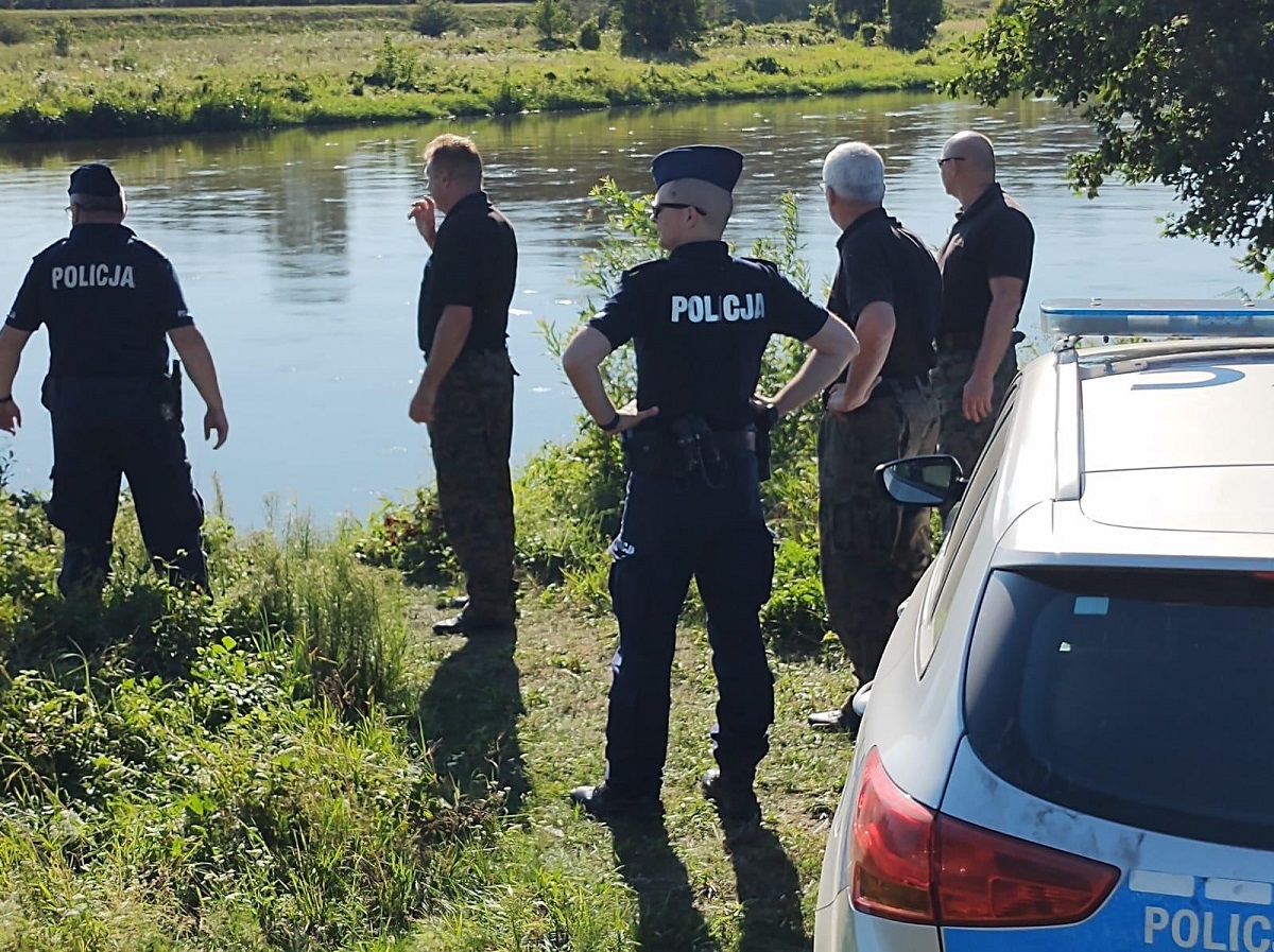 Nad rzeką, przy brzegu stoją umundurowani policjanci oraz funkcjonariusze Państwowej Straży Rybackiej.