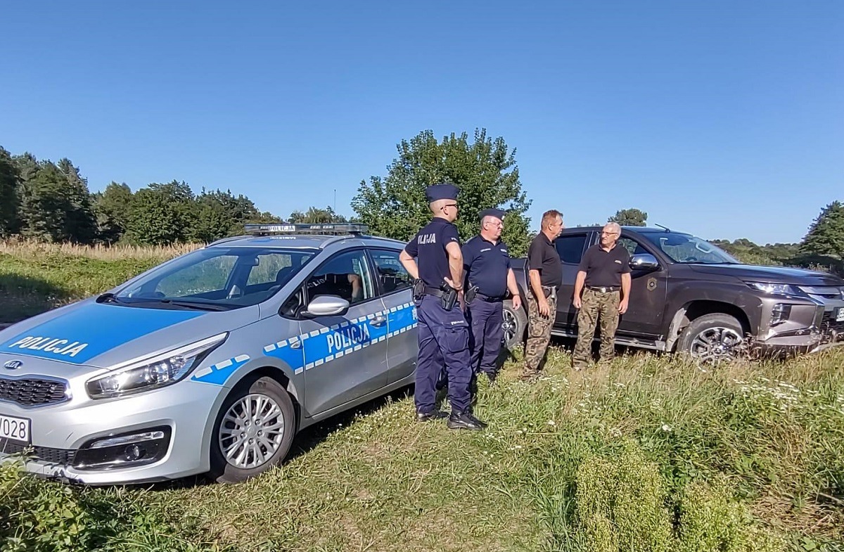 Na trawie, po lewej stronie stoi radiowóz policyjny, po prawo nieoznakowany samochód. Między pojazdami stoją umundurowani policjanci oraz funkcjonariusze Państwowej Straży Rybackiej.