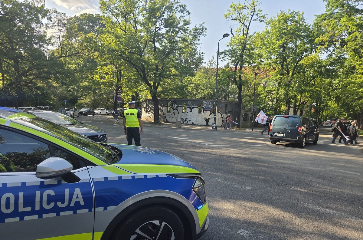 Radiowóz, za nim skrzyżowanie na którym stoi policjant, w tle przejście dla pieszych, przez które przechodzą osoby z flagami.