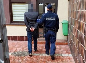 Policjant ubrany w niebieski mundur prowadzi zatrzymanego przez dziedziniec prokuratury