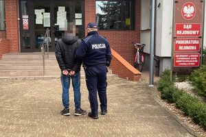 Policjant ubrany w granatowy mundur idzie z zatrzymanym do prokuratury. Stoją na chodniku przed schodami prowadzącymi do budynku prokuratury.