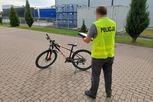 Policjant w cywilnym ubraniu, ma założoną żółtą kamizelkę odblaskową z napisem policja i dokonuj oględzin skradzionego roweru.