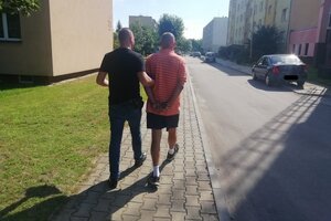 Policjant prowadzi mężczyznę, który ma założone kajdanki, zdjęcie zrobione od tyłu.