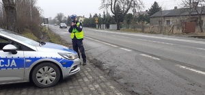 Na zdjęciu policjant drogówki, stoi koło radiowozu i mierzy prędkość kierujących pojazdami.