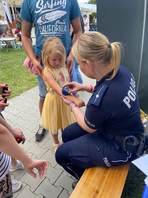 Policjantka na przedramieniu dziewczynki przystawia pieczątkę, wokół grupa dzieci