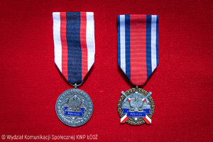 na czerwonym tle leżą dwa medale: Medal &quot;Zasłużony dla Policji&quot; oraz odznaczenie &quot;Zasłużony Policjant&quot;.