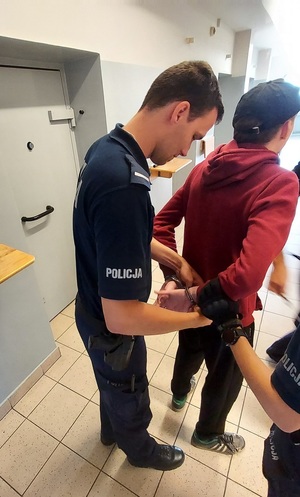 Policjant w pomieszczeniu PDOZ zakłada kajdanki zatrzymanemu.