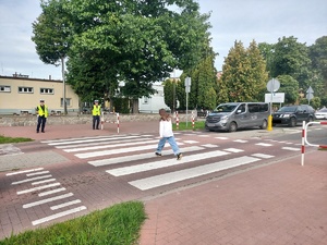 Przejście dla pieszych. Po drugiej stronie, przy pasach stoją policjanci, przez przejście przechodzi dziecko, samochody zatrzymują się przed pasami.