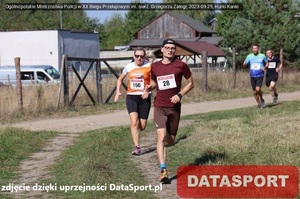 Ubita drogą, biegnie Arkadiusz Maruszewski, w dalszym planie biegną inni uczestnicy biegu. W prawym dolnym roku, na czerwonym tle jest napis Datasport. Po lewej stronie jest napis: zdjęcie dzięki uprzejmości DataSport.pl