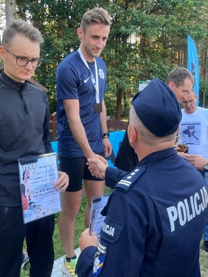 Po lewej stronie Arkadiusz Maruszewski na podium, stoi z dyplomem, stojący na wprost niego policjant w mundurze wręcza dyplom zwycięscy na I miejscu.