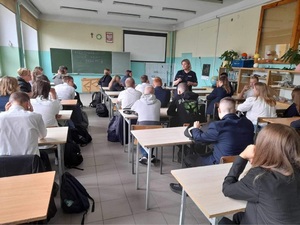 Szkolna klasa, uczniowie siedzą w ławkach, w klasie jest policjantka, która rozmawia z zebranymi.