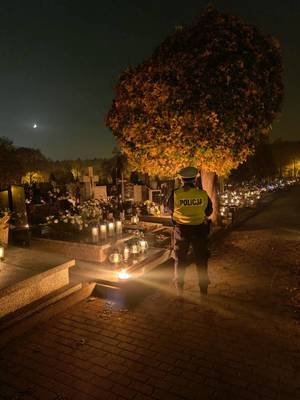Policjant na cmentarzu, jest ciemno, widać na grobach palące się znicze.