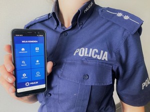 Umundurowany policjant trzyma w dłoni telefon z włączona aplikacją &quot;Moja Komenda&quot;. Telefon jest odwrócony przodem do zdjęcia. Widać stronę aplikacji na wyświetlaczu telefonu.
