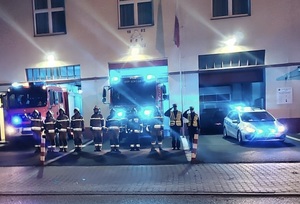 Przed budynkiem OSP, stoją od lewej strony strażacy, a po ich prawej stronie policjanci. Za policjantami i strażakami, stoją pojazdy służbowe z włączonymi sygnałami świetlnymi.