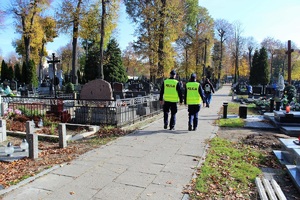 Policjant i policjantka ubrani w kamizelki, na plecach mają napis Policja. Idą alejką na cmentarzu.