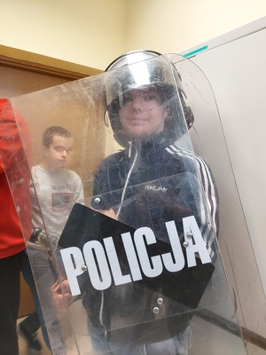 Uczeń ma założony hełm i trzyma tarczę z napisem Policja.
