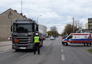 Ulica w mieście, po lewej stronie stoi policjant, przed pojazdem ciężarowym, po prawej karetka pogotowia.