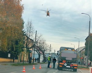 Ulica w mieście, po prawej stronie stoi pojazd ciężarowy, policjant rozmawia z mężczyzną. w tle widać wznoszący się helikopter ratowniczy.
