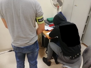 Osoba zatrzymana ma czarna bluzę z kapturem, który jest założony na głowę, podpisuje dokumenty, po lewej stronie stoi ubrany po cywilnemu policjant, ma na prawym przedramieniu opaskę z napisem policja.