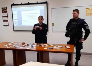 Policjantka i policjant w mundurach, stoją przed stołem, na którym są rozłożone przedmioty spakowane w małe strunowe torebki. Policjantka trzyma jeden przedmiot w dłoni, mówi.