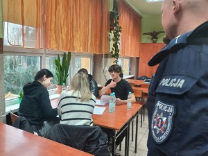 Uczniowie siedzą przy stole, rozwiązują wspólnie test, jeden z uczniów podnosi kciuk w geście ok, policjant stoi w pobliżu, przygląda się im.